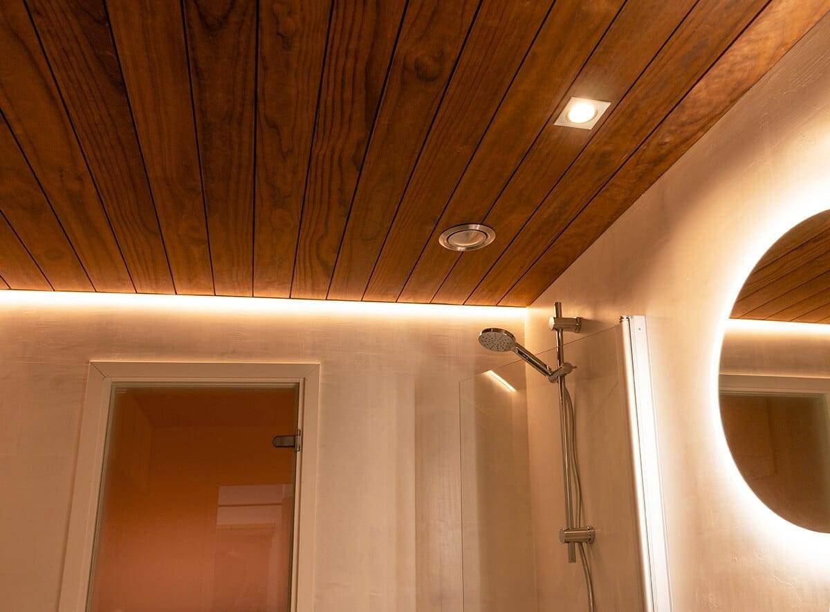 Modern bathroom with wooden panel ceiling, recessed LED lighting, round backlit mirror, metal showerhead, and frosted glass door ajar in the background.
