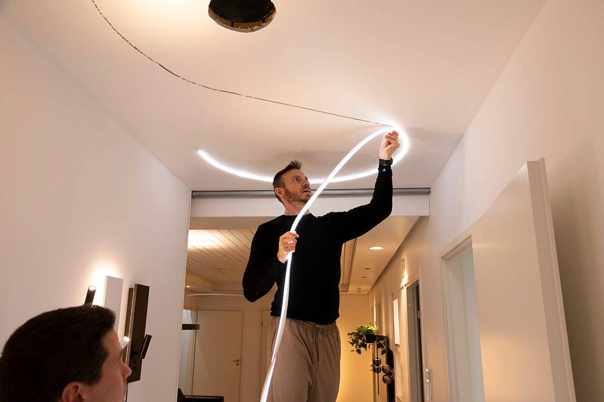 A person attaches a flexible LED strip light to a modern ceiling, which improves the lighting in the room. Black sweater, beige pants; another person in foreground, blurred.