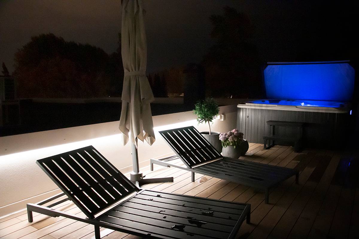 Two lounge chairs and a closed parasol on the terrace in the evening twilight. LedStore.fi's high quality LED courtyard lighting softly illuminates the glowing, blue-tinted hot tub in the background of the courtyard of this modern house.