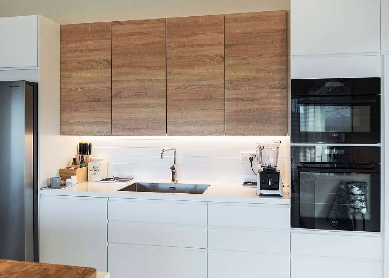 Modern kitchen with sleek LED lighting, light wood cabinets, white countertop, built-in oven & microwave, stainless steel fridge; knife block & blender.