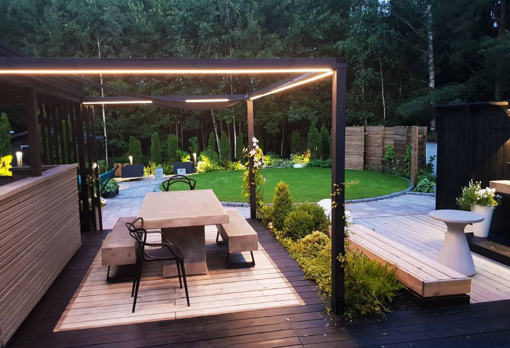 Modern outdoor terrace with pergola with built-in LED lighting, concrete table, benches with wooden deck; lush garden and manicured lawn area.