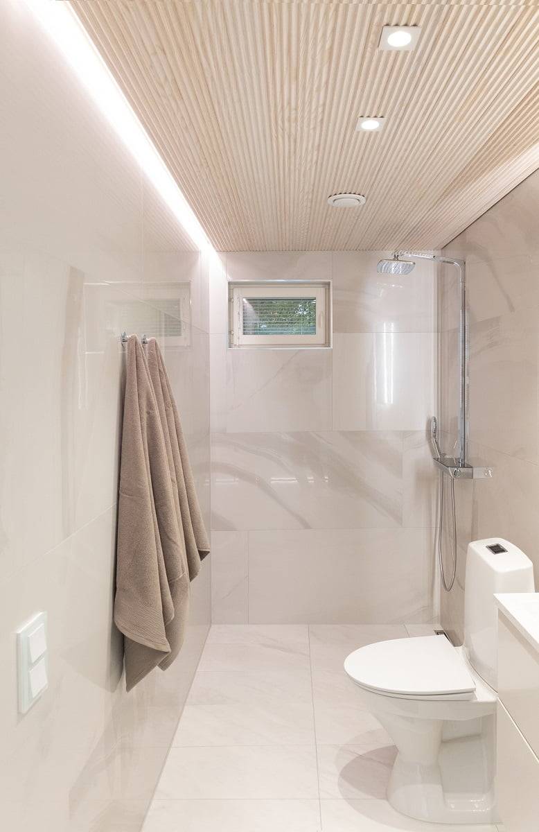 A modern and minimalist bathroom with light beige walls and a wooden tiled ceiling. A wall-mounted towel rail with two towels, a small window and a shower area separated by a glass partition.   