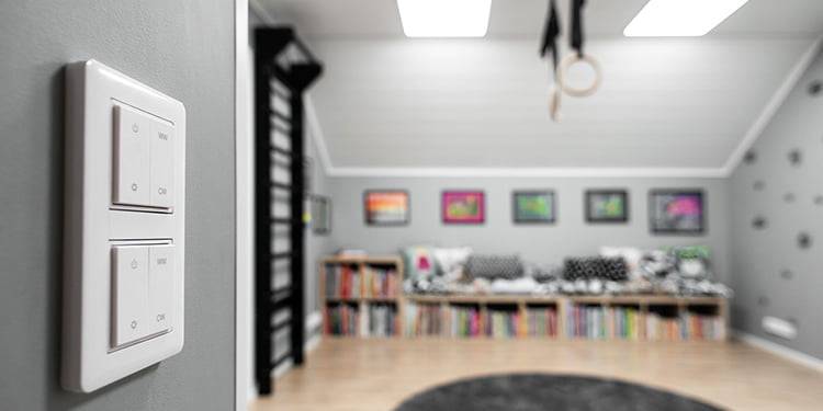 Modern room with light gray walls, double light switch foreground; cozy seating area with colorful frames, bookshelves, circular rug; warm LED ceiling lights.