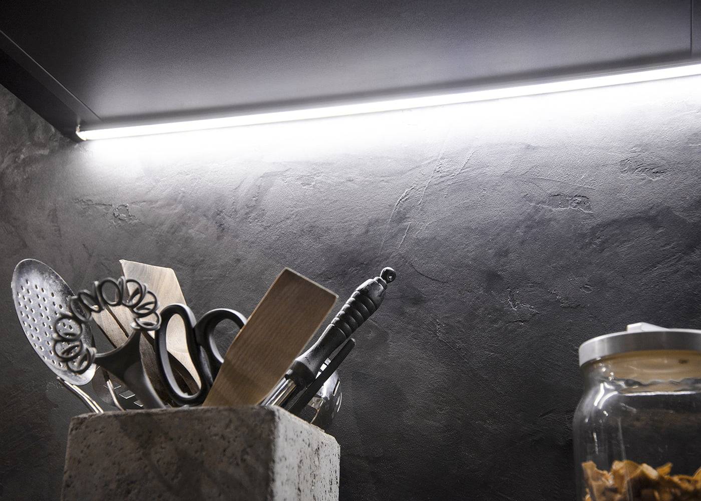 Kitchen utensils in a stone holder under bright LED lighting, featuring a whisk, spatula, and scissors on a textured countertop with a glass jar nearby.