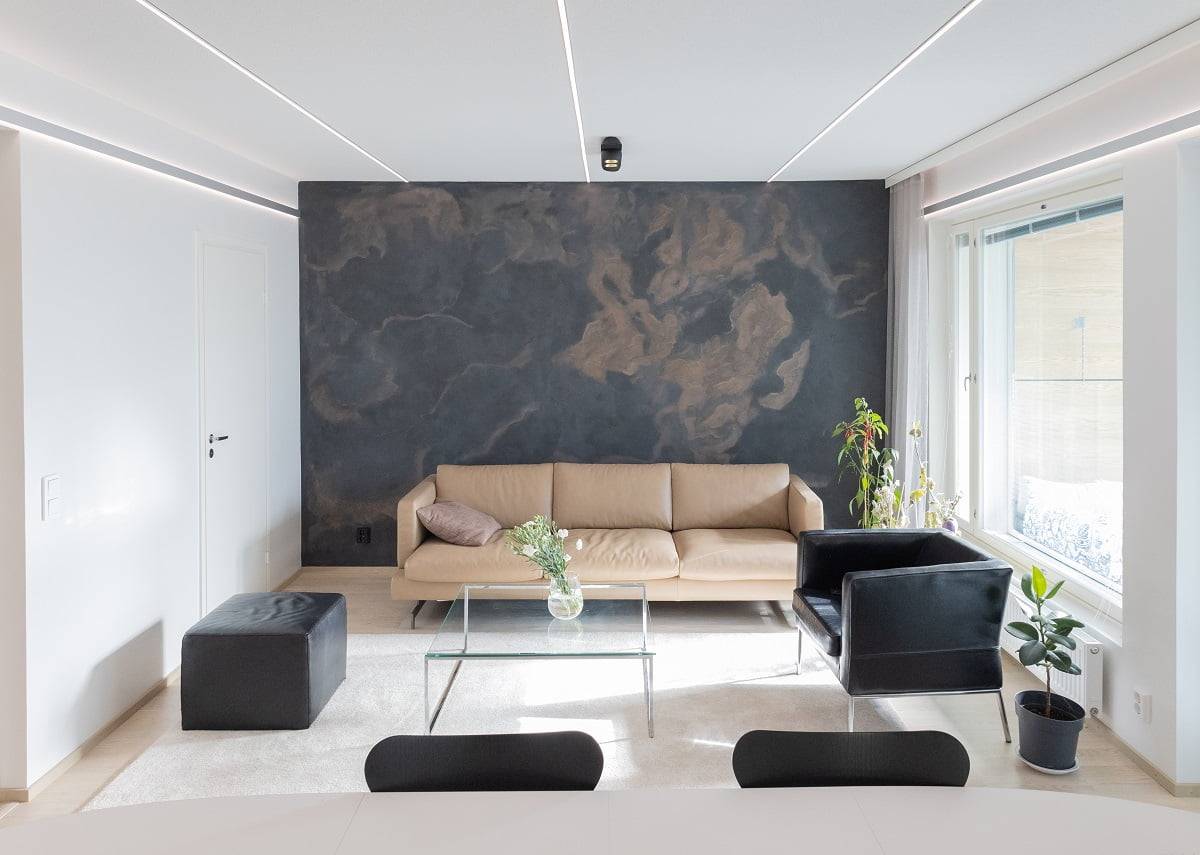 The modern living room has a beige sofa, black armchair and glass coffee table, which match the dark effect wall. Next to the window stands a green plant near a small black straw. Order a spotlight, complemented by wild LED lights from LedStore.fi.