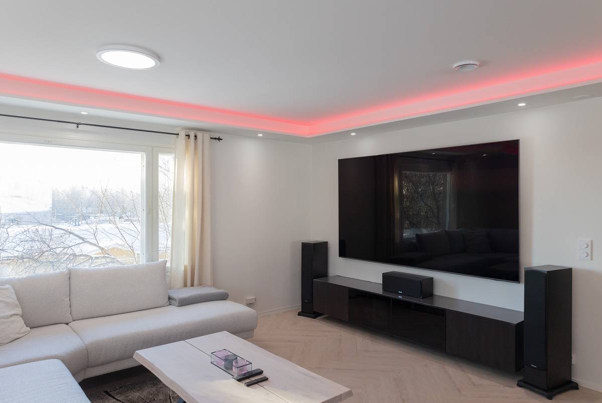A modern living room full of elegant atmosphere. A large sofa invites you to relax, while a light wooden coffee table adds warmth to the space. A wall-mounted flat-screen TV with LED backlighting adds a modern touch to the room. Amplify your experience with floor speakers and enjoy the red LED accent lighting on the ceiling. A large window lets in natural light, making the space feel spacious and bright. All these elements combine to create the perfect environment with LedStore.fi's versatile LED lighting solutions.