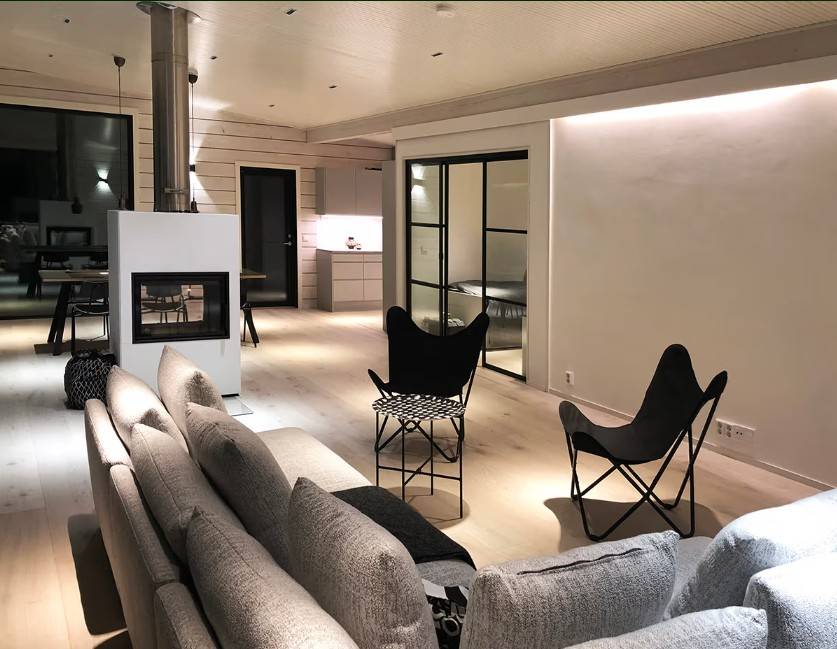 A modern living room with a grey sofa, black chairs, a fireplace and an open-plan kitchen in the background - the atmosphere and style is created by LedStore.fi's high-quality LED lighting solution.