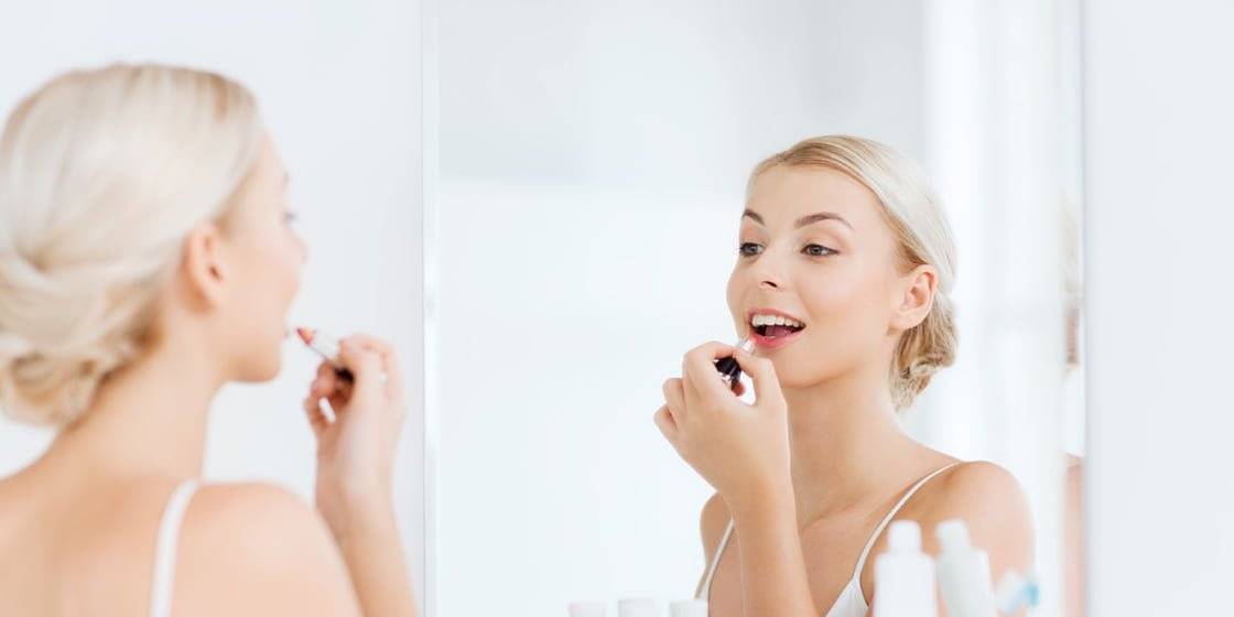 Woman with blonde hair applying lipstick in front of mirror lit by LED lights in bathroom; white top, toiletries visible, bright and soft lighting.