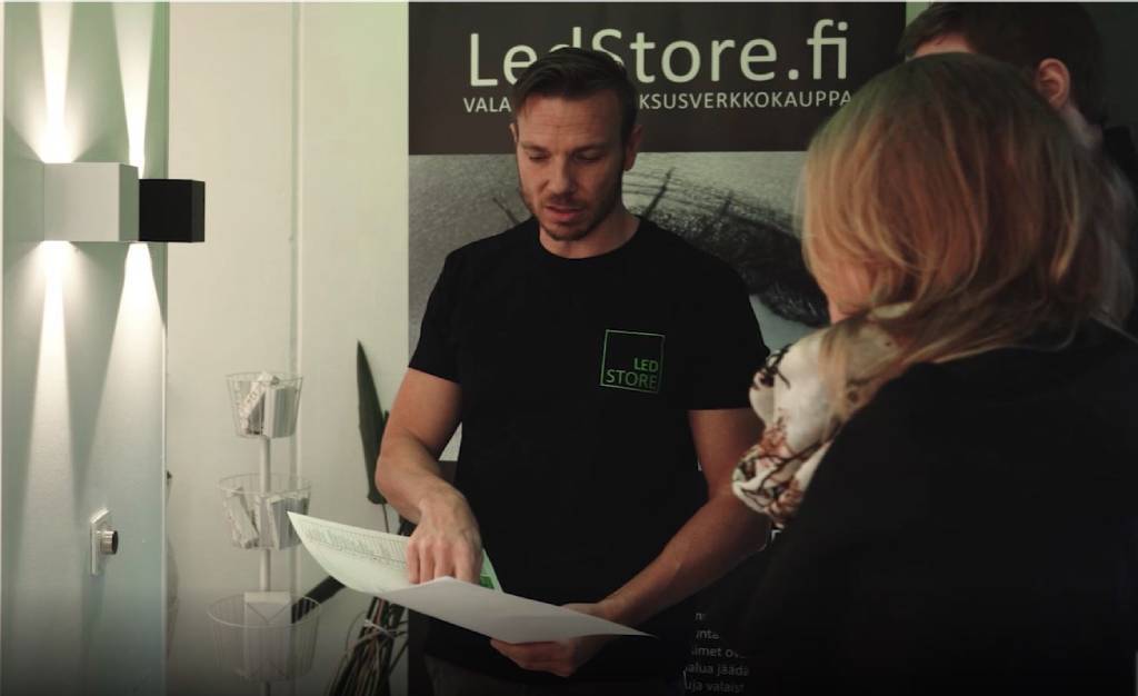 Man in LedStore.fi shirt discusses LED document with blonde woman in a store showcasing various lighting fixtures; third person partially visible.