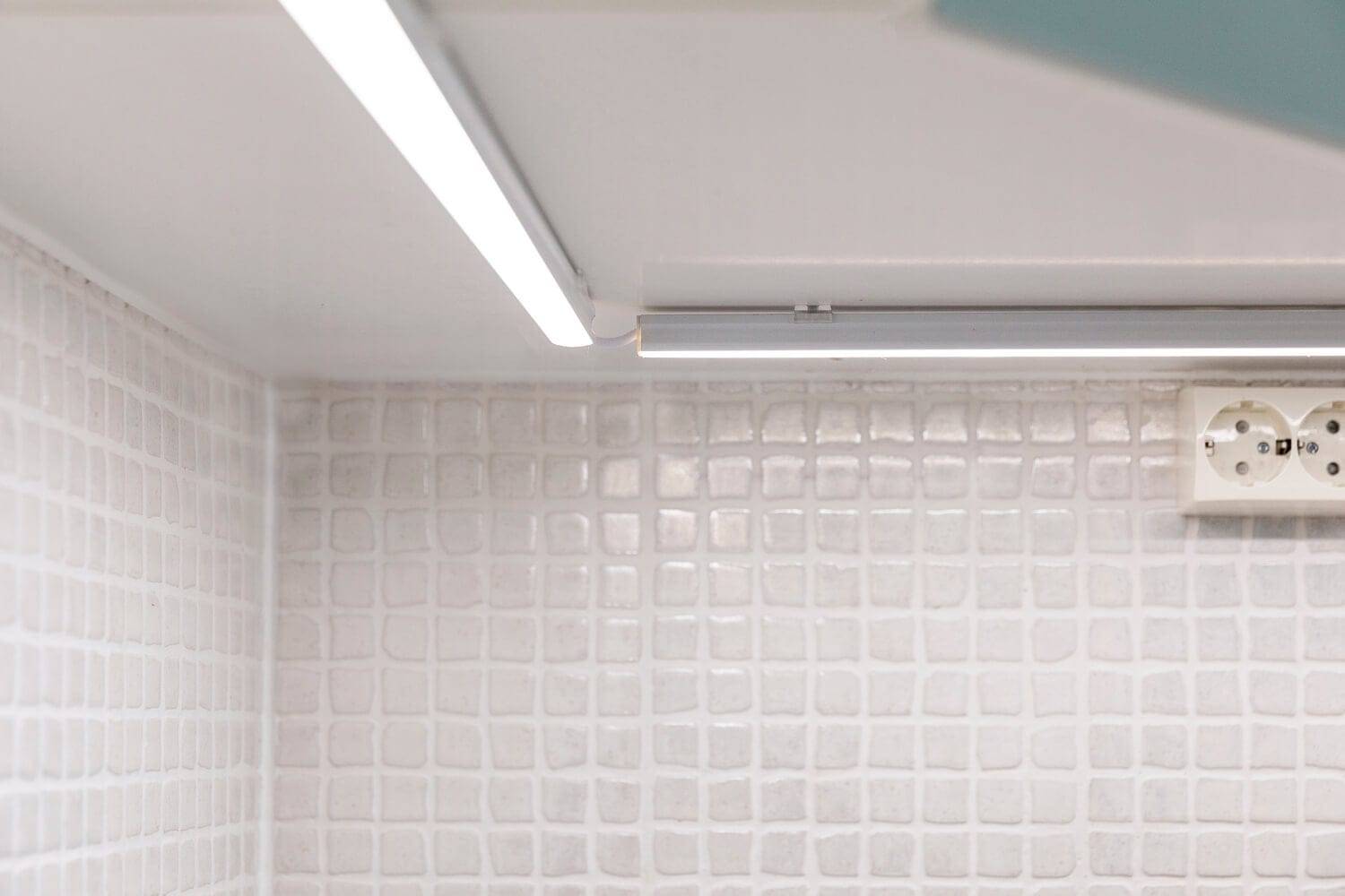 A modern kitchen backsplash with white textured tiles illuminated by a linear LED light under the cabinet; a gentle glow highlights the wall socket.