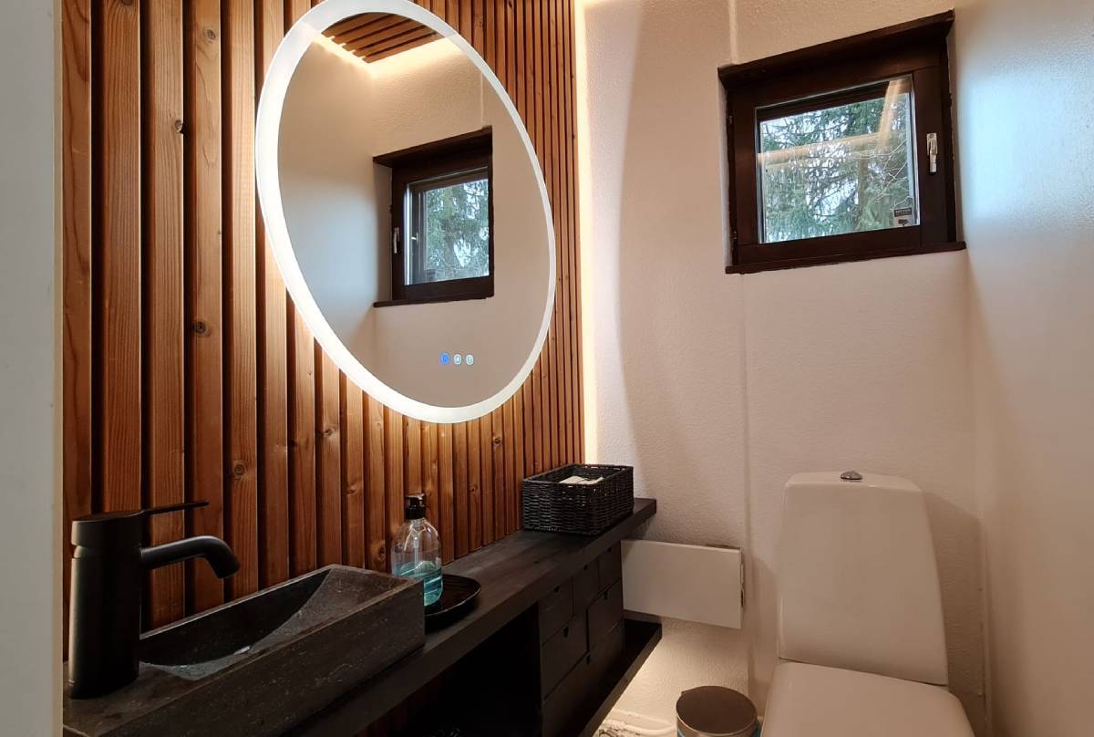 A modern bathroom with LED lighting provided by LedStore.fi includes a circular LED mirror on top of a wood panel wall. Below the mirror is a black countertop with a rectangular sink and black faucet and soap dispenser. A small window above the toilet seat, and a handy storage solution is provided nearby.