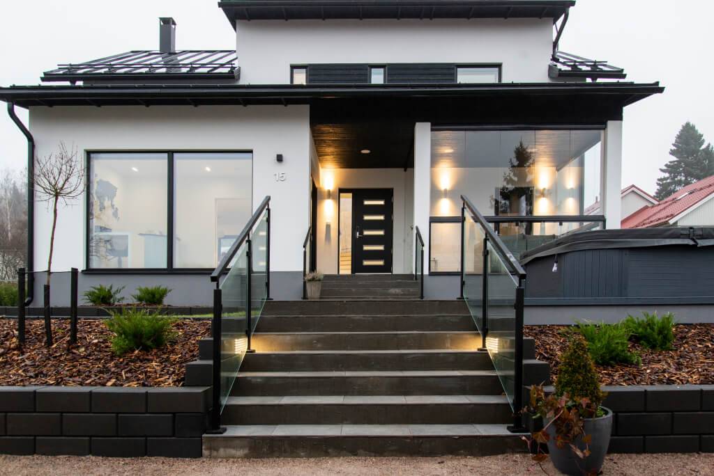 Modern house with white façade and black accents; stairs with glass balustrades and LED lights. Large windows, minimal landscaping, shrubs and neighbouring house.