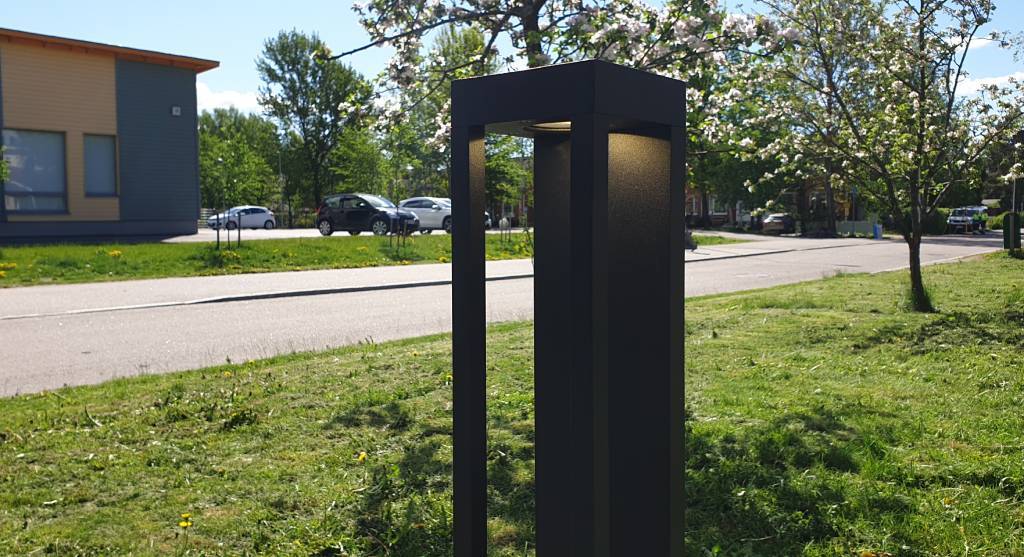 Modern black LED outdoor light fixture on grassy lawn; trees with blooming branches and building in background on sunny day.