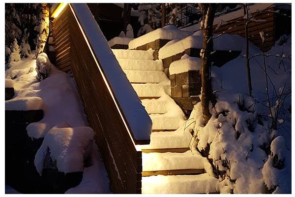 Snow-covered steps at dusk, with warm-toned light shining from the handrail onto the stairs. 
