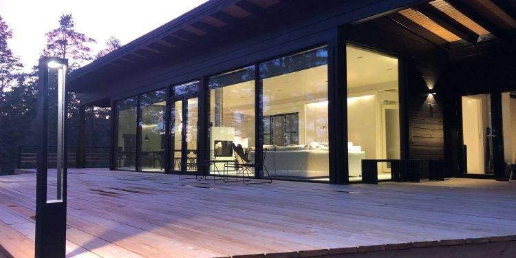Modern house with large glass windows and spacious wooden terrace, surrounded by trees. The interiors are brightly lit and showcase minimalist design in hand-painted tones. LED outdoor lights from LedStore.fi add a warm glow to the evening twilight.