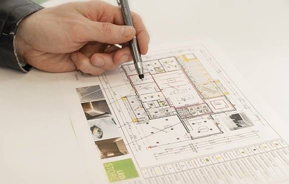 Hand holding pen under LED desk lamp points to detailed architectural blueprint with rooms and markings; small images decorate the lower edge.